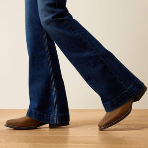 A close-up of a person's legs wearing dark blue flared jeans and brown suede boots, standing on a light wood floor against a plain beige wall.