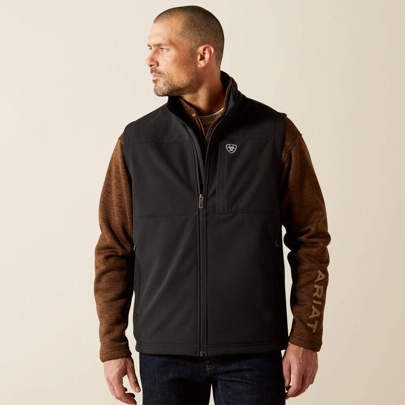 A man wearing a black zip-front vest with brown sleeves and dark blue jeans, photographed against a plain light beige studio background. The vest has a small shield logo on the chest and 'ARIAT' text printed vertically on one sleeve.