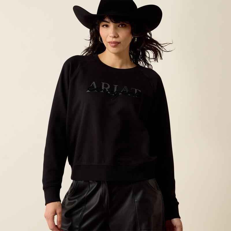 A woman wearing a black crewneck sweatshirt with a tonal black 'ARIAT' logo on the chest.