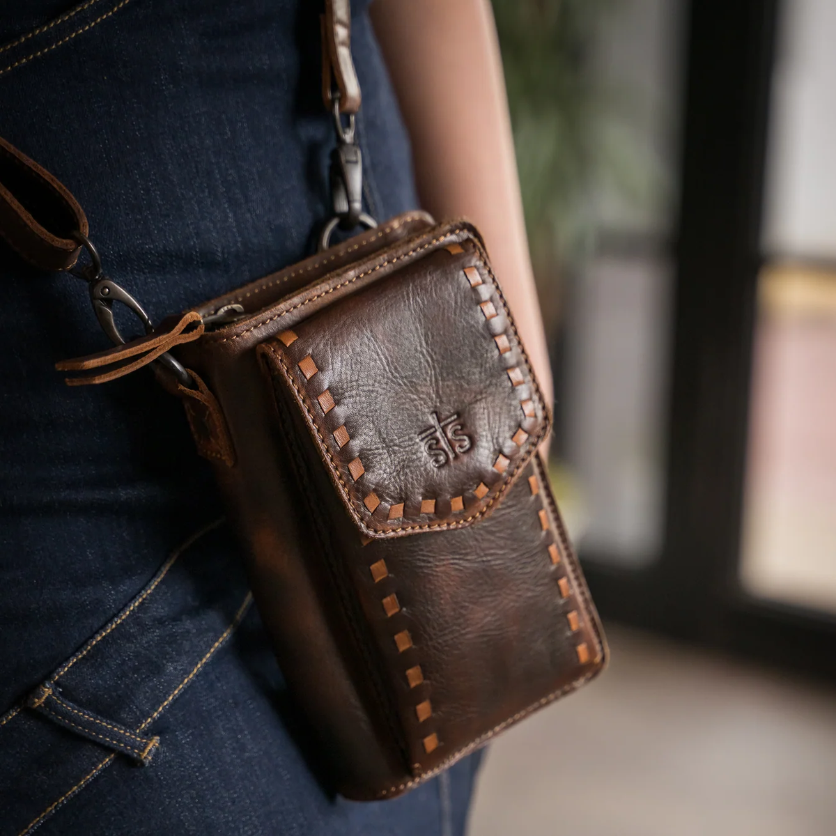 A brown leather crossbody phone pouch with decorative stitching and an embossed logo, worn crossbody over dark blue denim jeans.