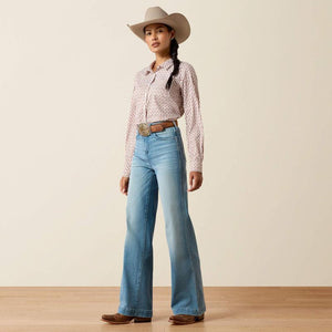 A woman modeling a western-style outfit featuring light blue wide-leg jeans, a pink and white patterned long-sleeve shirt, a brown leather belt with a large silver buckle, a beige cowboy hat, and brown boots, set against a plain beige studio background.