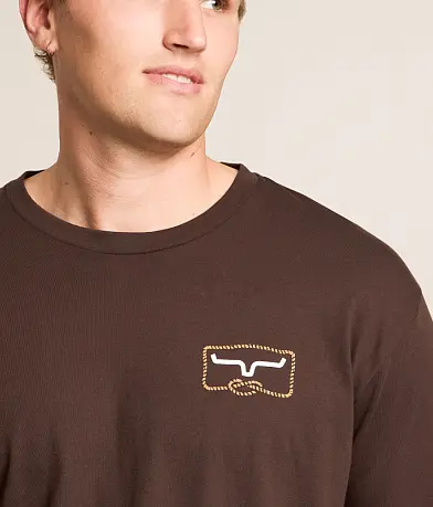 A close-up of a man wearing a dark brown ribbed long-sleeve t-shirt with a small embroidered logo on the left chest.
