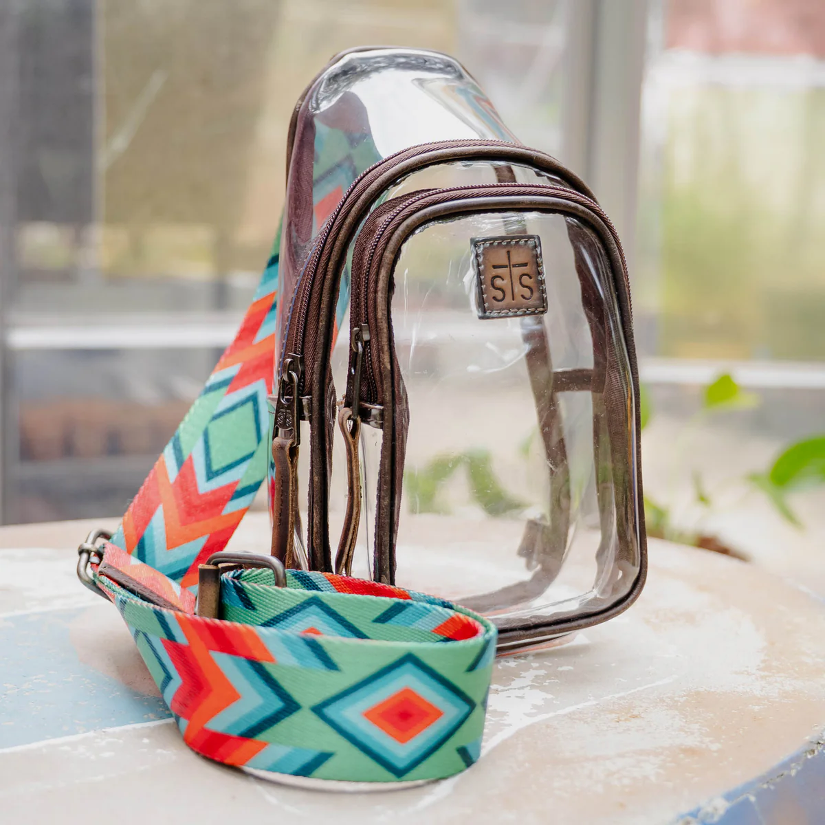 Clear plastic bag with brown strap and colorful geometric patterned strap on a surface.