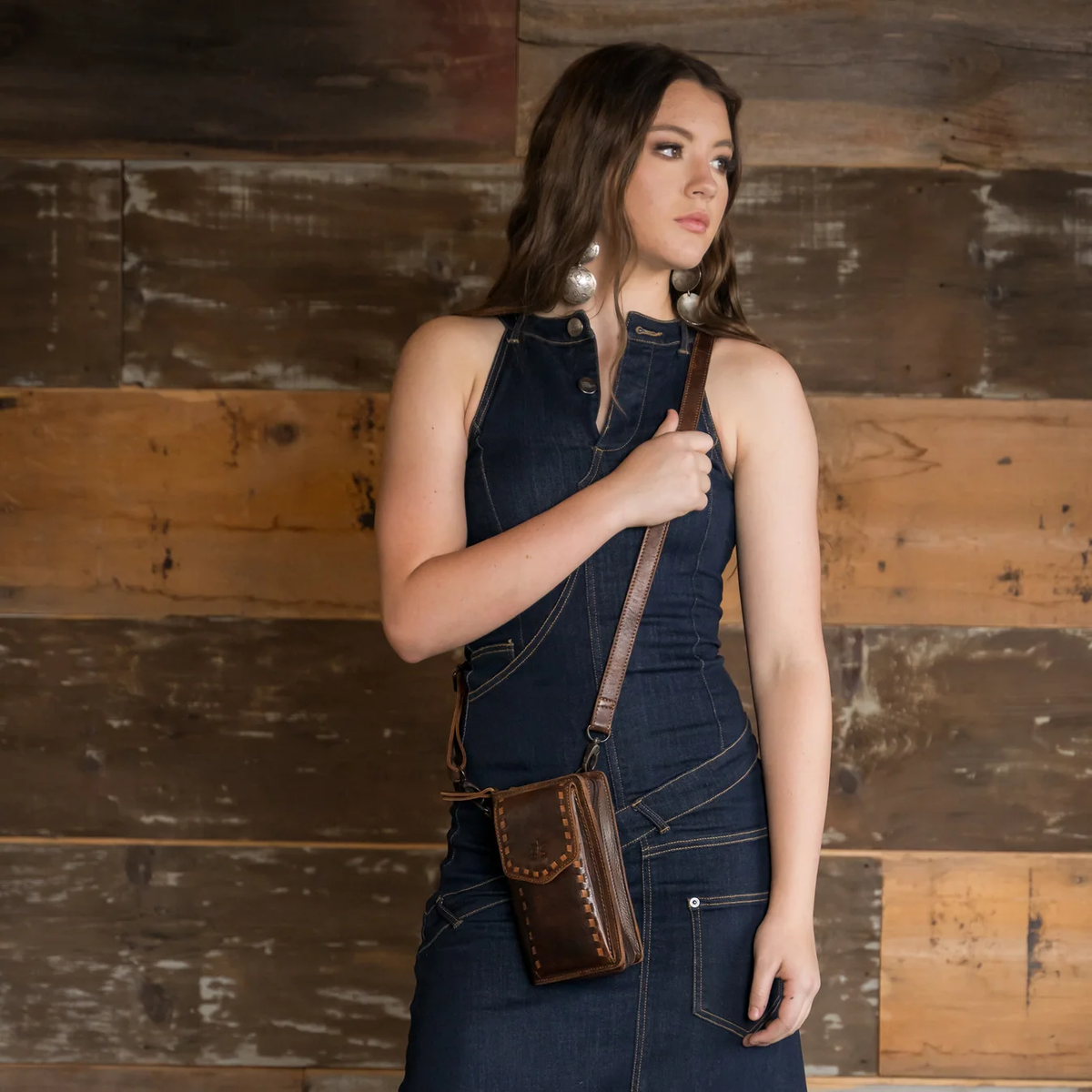 A woman wearing a dark denim overall dress stands against a rustic wooden plank wall, carrying a brown leather crossbody pouch bag with a matching strap.