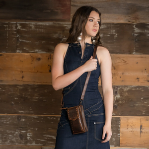 A woman wearing a dark denim overall dress stands against a rustic wooden plank wall, carrying a brown leather crossbody pouch bag with a matching strap.