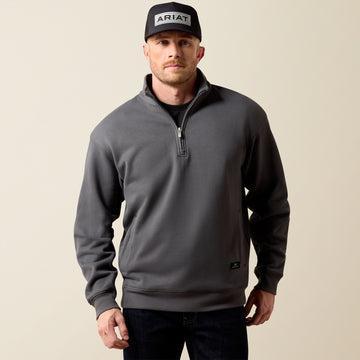 A man wearing a dark gray quarter-zip sweatshirt, a black cap with 'ARIAT' on the front, and dark jeans, standing against a solid beige background.