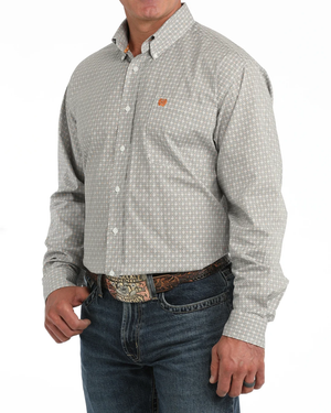A man wearing a liwhite micro-patterned button-down shirt with a small orange logo on the chest, tucked into blue jeans with a western belt.