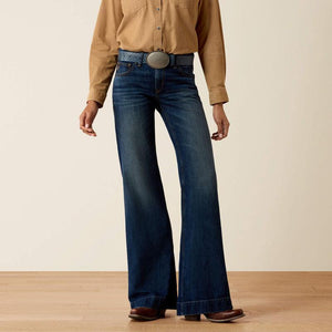 A person wearing dark blue wide-leg jeans with a tan button-up shirt, a blue belt with a large silver buckle, and brown shoes, standing against a plain beige wall and light wood floor.