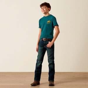 Young boy wearing a teal t-shirt and dark jeans standing against a plain background