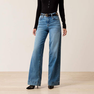 A model wearing high-waisted, wide-leg blue denim jeans with a black long-sleeve top and black pointed-toe heels, standing against a neutral beige studio background.