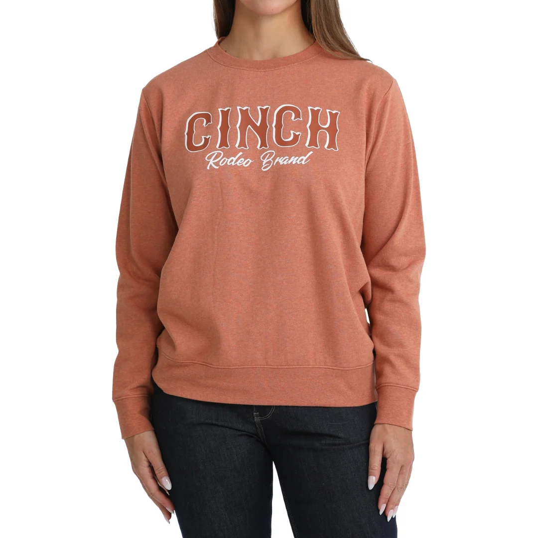 A woman wearing a heathered terracotta crewneck sweatshirt with 'CINCH Rideos Brand' printed in white and brown outline across the chest, paired with dark jeans, against a plain white background.