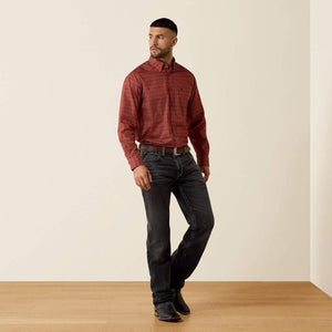 A man modeling a long-sleeve button-down shirt with a subtle red and black textured pattern, paired with dark gray jeans and black boots, standing against a minimalist beige wall and light wood floor.