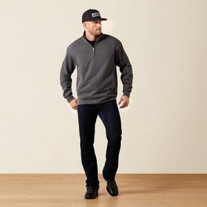 A man wearing a dark gray quarter-zip pullover sweatshirt, dark blue jeans, and a black baseball cap stands against a plain beige studio wall with a light wood floor.