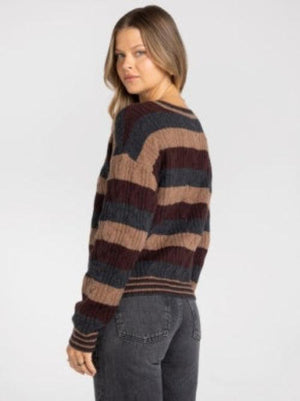 A woman wearing a long-sleeve crewneck sweater with horizontal stripes in dark brown, tan, and charcoal gray