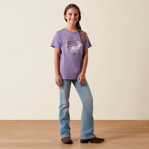 Young girl wearing a purple t-shirt and blue jeans standing on a wooden floor with a beige wall background.