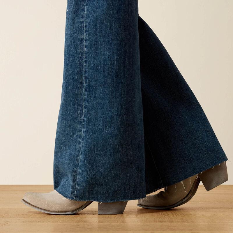 Close-up of a person's legs wearing dark blue flared denim jeans and taupe-brown western-style ankle boots, standing on a light wood floor against a beige wall.