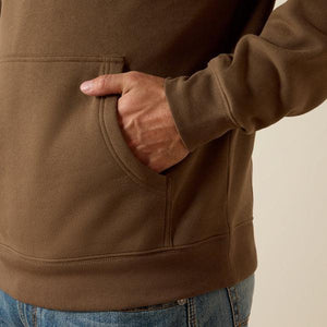 Close-up of a person wearing a brown pullover hoodie with a front kangaroo pocket, shown with one hand in the pocket against a plain light beige background.