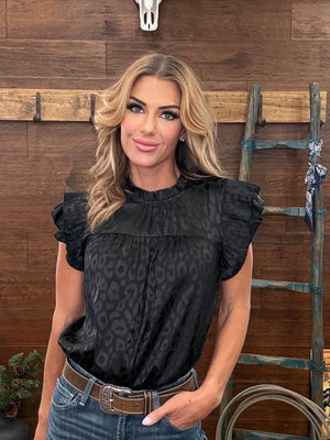 Woman wearing a black patterned top and blue jeans against a wooden wall.