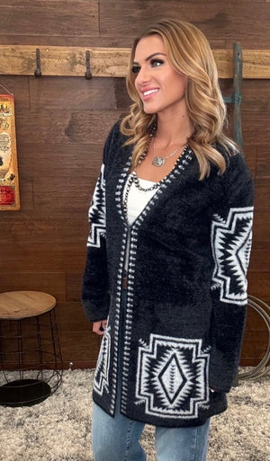 A woman wearing a black fuzzy cardigan with white geometric patterns, styled with a white top and blue jeans, standing in front of a rustic wooden wall.