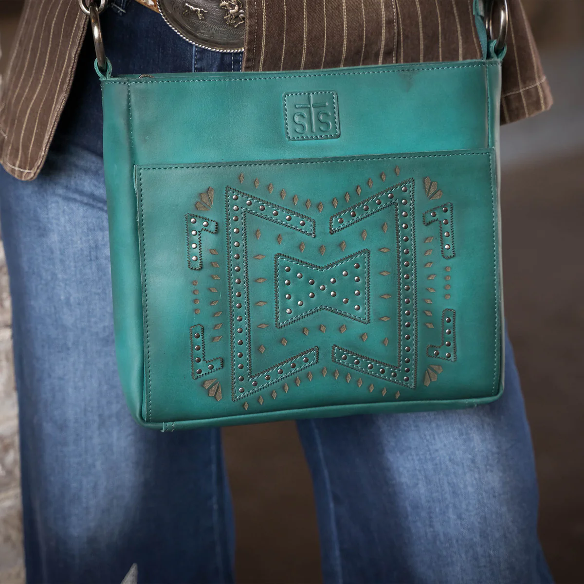 Turquoise handbag with decorative patterns held by a person wearing jeans and a brown jacket.