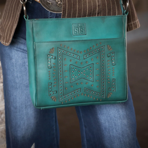 Turquoise handbag with decorative patterns held by a person wearing jeans and a brown jacket.