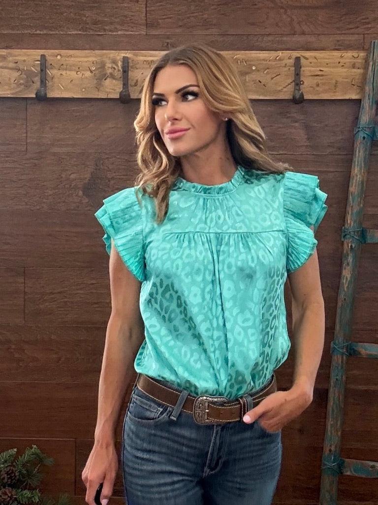 Woman wearing a turquoise blouse with ruffled sleeves and blue jeans against a wooden background