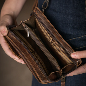 A close-up of a person holding an open brown leather wallet or clutch with multiple zip compartments and card slots, shown against dark denim clothing.