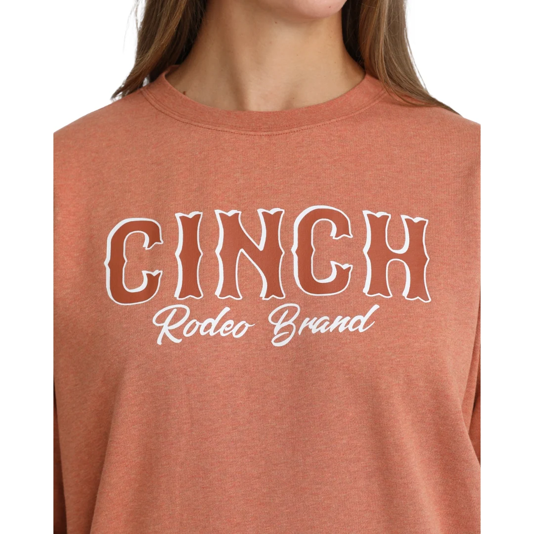 A close-up of a person wearing a heathered terracotta crewneck sweatshirt with 'CINCH Rodeo Brand' printed in white across the chest.