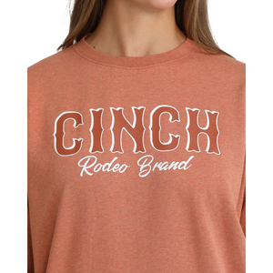 A close-up of a person wearing a heathered terracotta crewneck sweatshirt with 'CINCH Rodeo Brand' printed in white across the chest.
