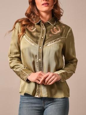 A woman wearing a long-sleeve western-style button-up shirt in olive green with embroidered details on the chest, paired with blue jeans, against a plain beige background.