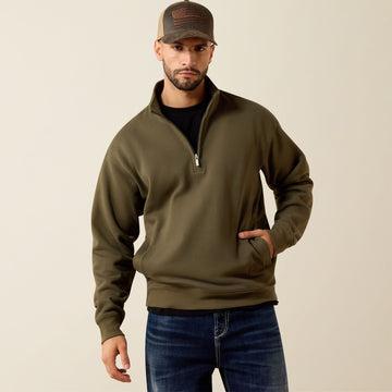 A man wearing an olive green quarter-zip pullover sweatshirt, blue jeans, and a brown baseball cap, standing against a plain beige studio background.
