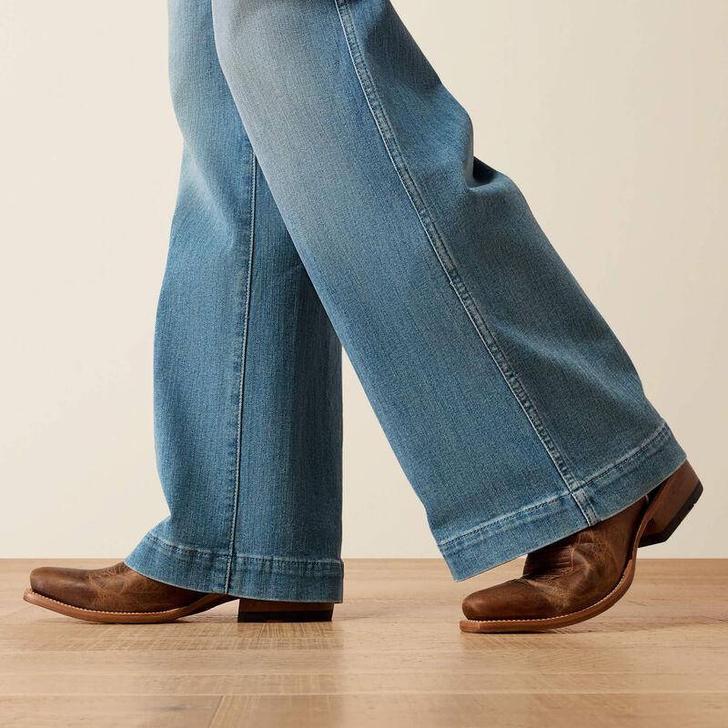 Close-up of a person wearing light blue wide-leg jeans and brown leather boots, standing on a light wood floor against a plain beige wall.