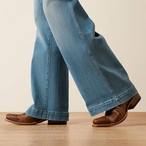 Close-up of a person wearing light blue wide-leg jeans and brown leather boots, standing on a light wood floor against a plain beige wall.