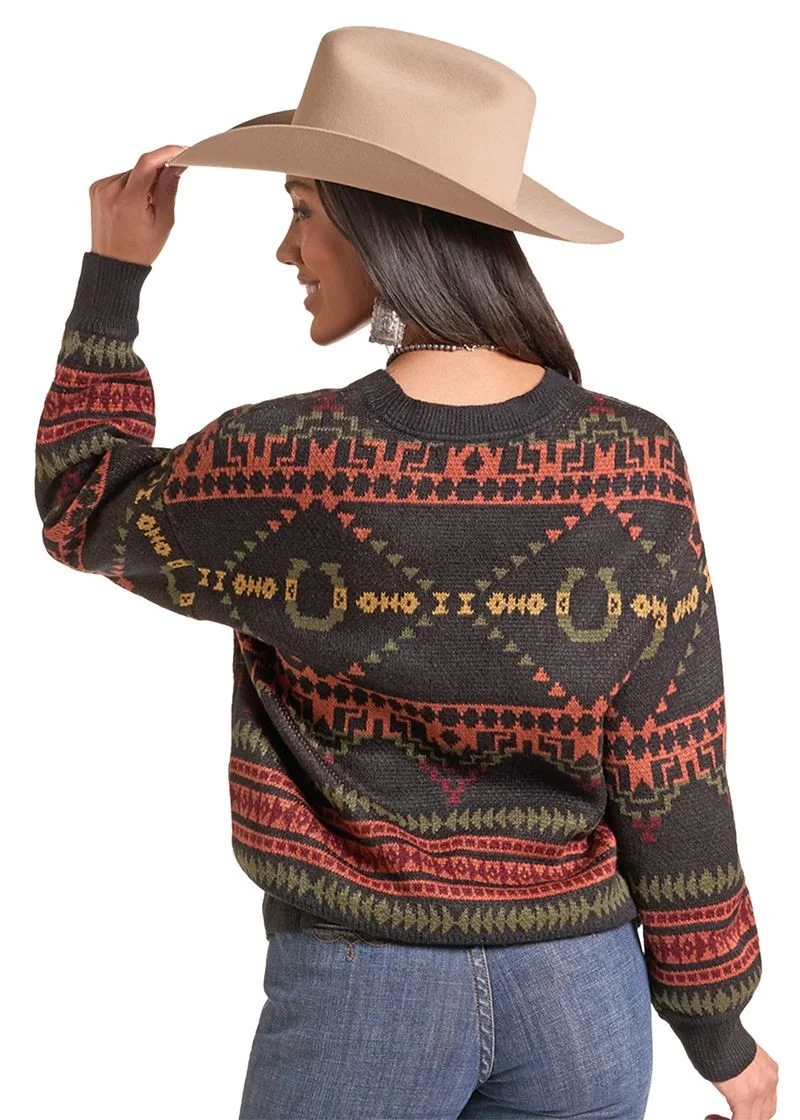A woman wearing a dark charcoal knit sweater with geometric and horseshoe patterns in rust red, olive green, and tan, paired with a beige cowboy hat and blue jeans, shown from a three-quarters back view against a white background.