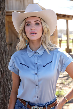 Woman wearing a light blue shirt with black arrows and a beige cowboy hat outdoors.