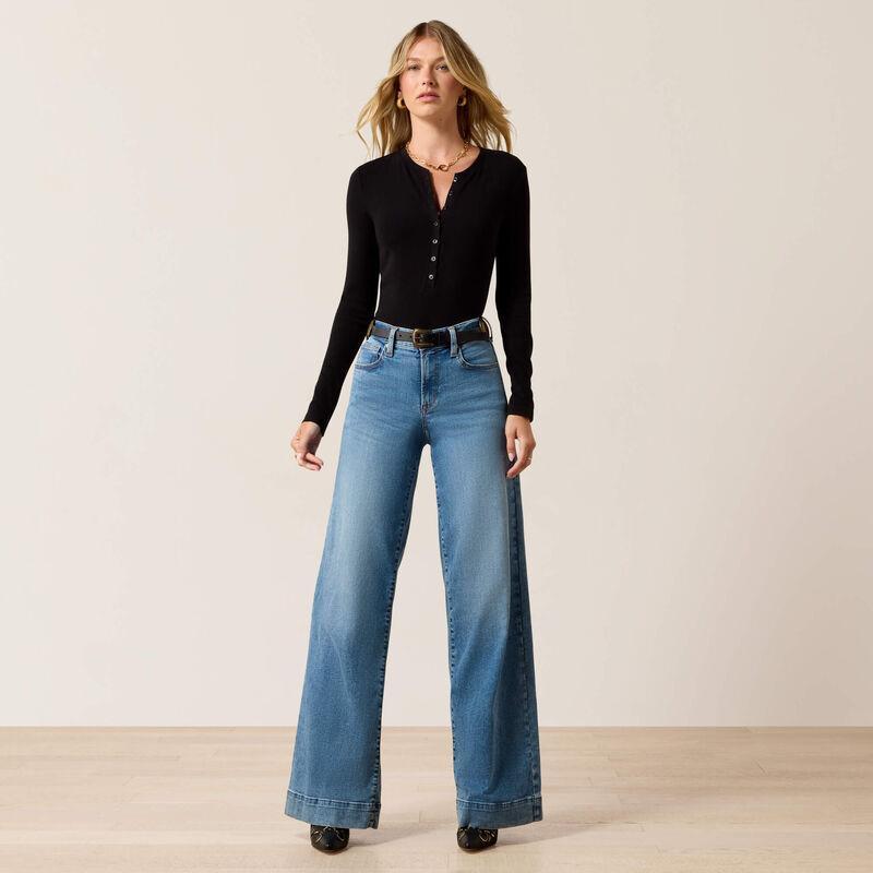 A woman modeling a black long-sleeve henley top and high-waisted, wide-leg blue jeans with a black belt, standing against a neutral beige studio background.