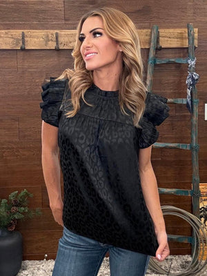 Woman wearing a black top with ruffled sleeves against a wooden wall.