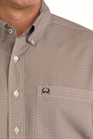 Close-up of a man wearing a brown and beige micro-patterned button-down shirt with a chest pocket and a small black embroidered logo.