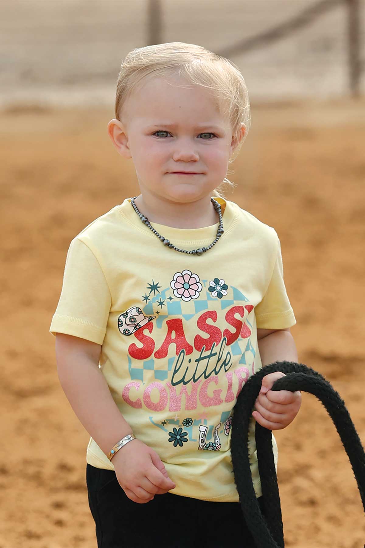 Child wearing a yellow shirt with colorful text and graphics, standing on a dirt surface.