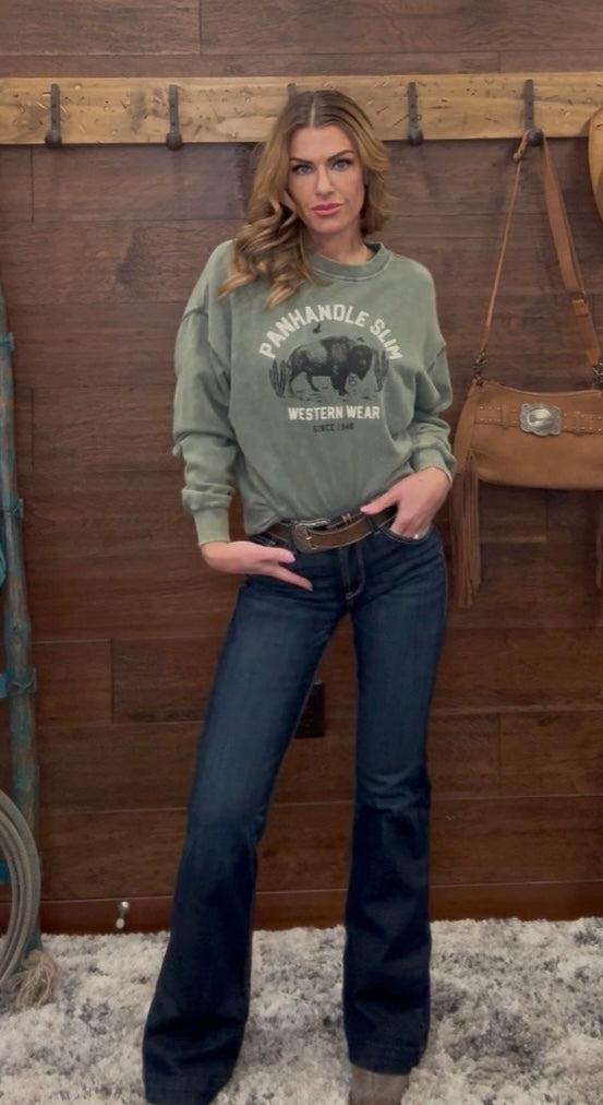 Woman wearing a green sweatshirt with a graphic design and dark jeans, standing against a wooden wall.