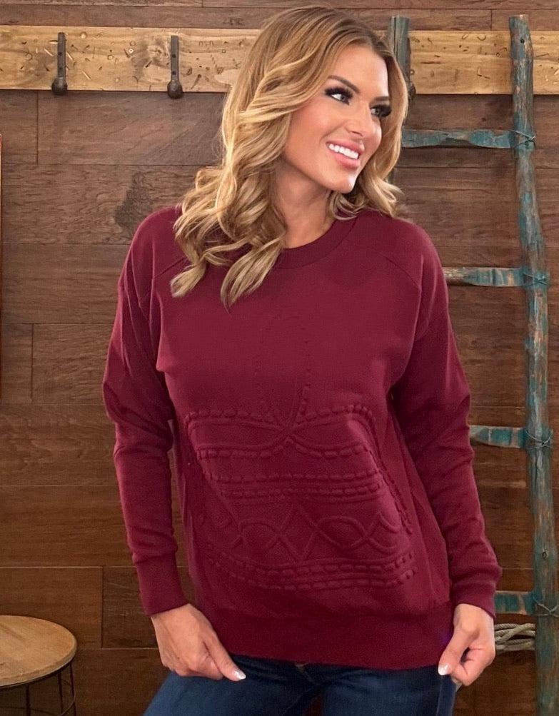 A woman wearing a burgundy long-sleeve knit sweater with a textured cable pattern on the front, paired with dark blue jeans and a brown belt, standing in front of a rustic wood-paneled wall.
