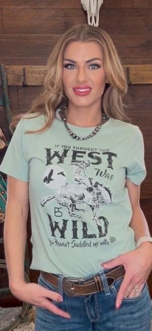 Woman wearing a light green t-shirt with a graphic design and text, standing against a wooden wall.