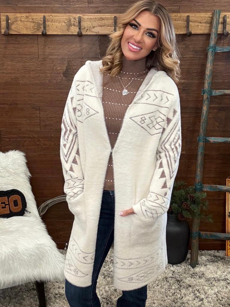 A woman wearing a long cream-colored fuzzy cardigan with brown geometric patterns, styled over a brown knit turtleneck sweater and dark blue jeans, standing in a rustic room with a wooden wall and rustic decor.