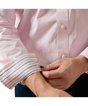 A close-up of a person wearing a light pink long-sleeve button-up shirt and adjusting the striped cuff, with dark blue jeans visible at the bottom.