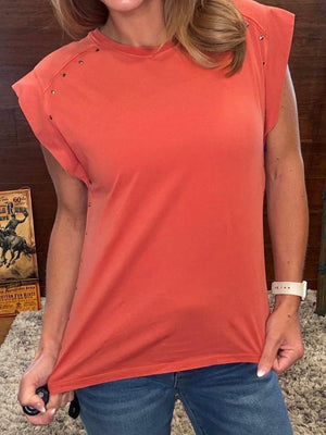 A woman wearing a coral orange sleeveless t-shirt with silver stud embellishments on the shoulders, paired with blue jeans.
