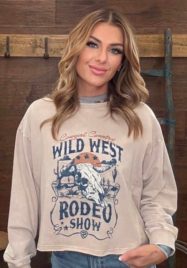 A woman wearing a cream-colored long-sleeve graphic sweatshirt with a 'Wild West Rodeo Show' design, posed against a rustic wooden wall.