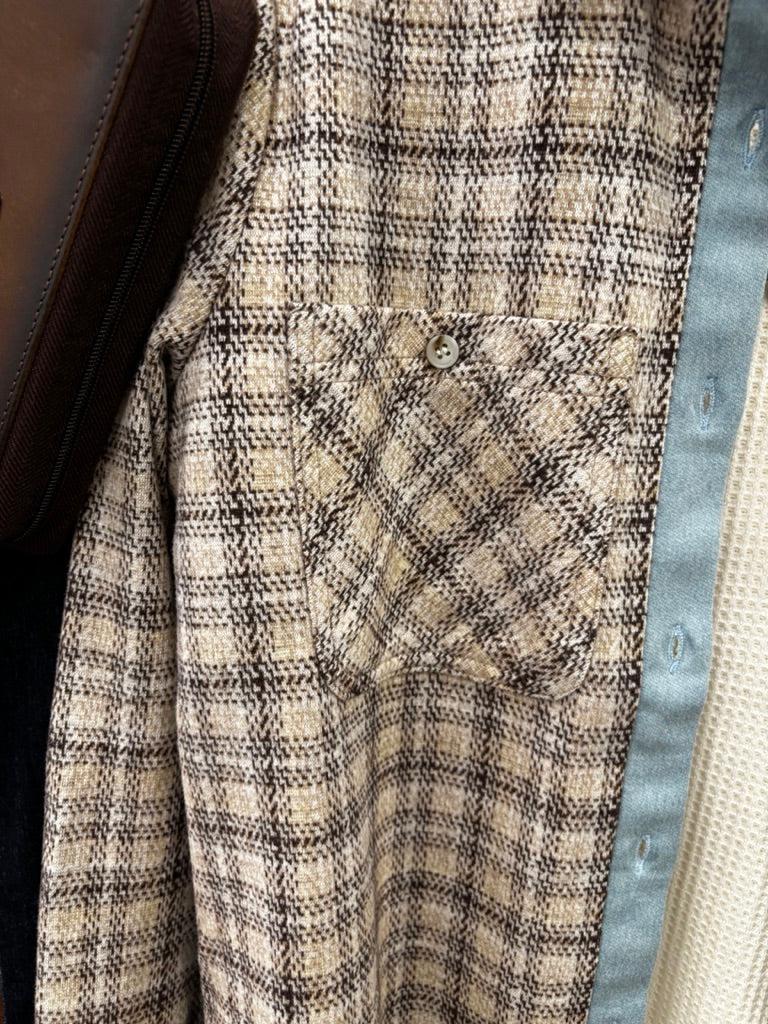 Close-up of a beige and brown plaid overshirt with a chest pocket, layered over a cream knit top and a light blue button placket.