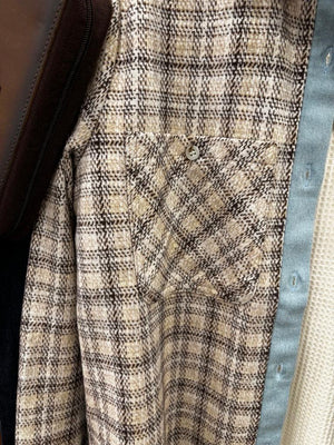 Close-up of a beige and brown plaid overshirt with a chest pocket, layered over a cream knit top and a light blue button placket.