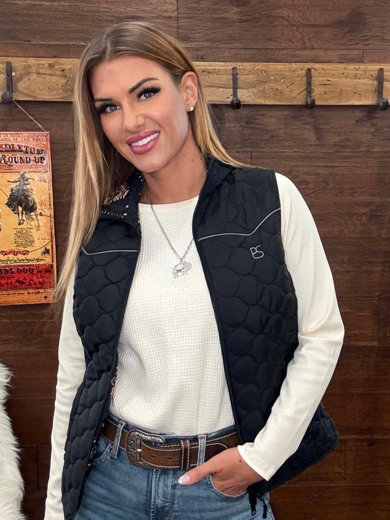 A woman wearing a black quilted sleeveless vest over a white long-sleeve top and blue jeans. 