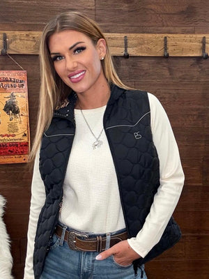 A woman wearing a black quilted sleeveless vest over a white long-sleeve top and blue jeans. 